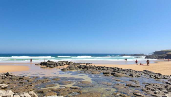 A sunlit beach featuring gentle waves, golden sands ideal for sunbathing, rocky formations for exploration, accessible ramps, and diverse individuals enjoying the day under a clear blue sky.